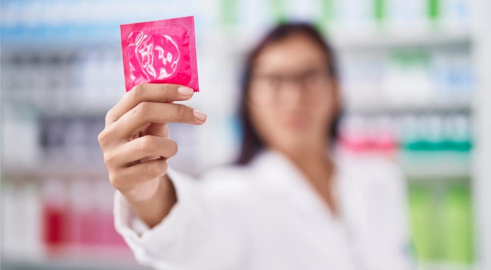 Jeune belle pharmacienne hispanique souriante confiante tenant un préservatif à la pharmacie